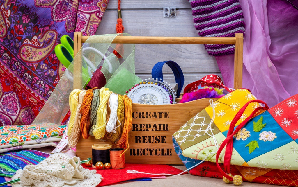 A colorful craft station with various fabrics, threads, scissors, and sewing tools arranged around a wooden box labeled "CREATE, REPAIR, REUSE, RECYCLE," emphasizing sustainable crafting and upcycling.