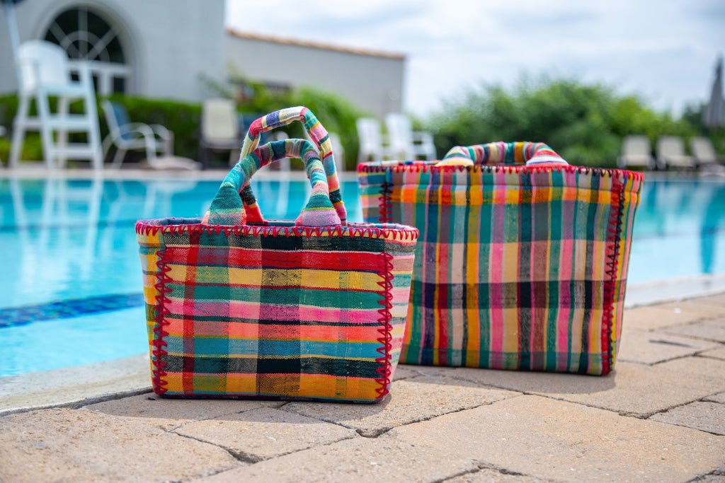 Two vibrant, handwoven tote bags with colorful plaid patterns placed beside a swimming pool under a sunny sky.