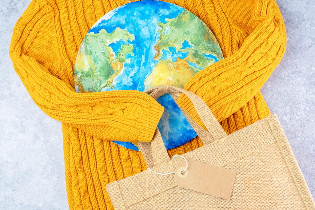 A sustainable lifestyle concept featuring a yellow knitted sweater, a reusable burlap tote bag with a blank tag, and a painted globe, symbolizing eco-conscious living and environmental responsibility.