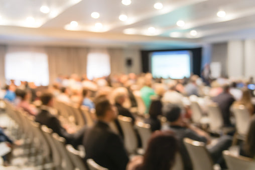 Blurry indoor view of engaged diverse professionals seated in rows at a well-lit conference venue, facing a large projector screen on stage amid elegant drapes and spotlights.
