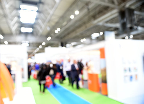 Blurry dynamic view of diverse attendees walking through a vibrant trade fair hall, featuring white exhibit booths with orange accents, blue carpet aisle on green flooring, and industrial ceiling lights.