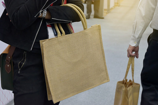 Stylish urban couple on sunny street: woman in black jacket and skirt holds large beige jute tote; man in white shirt carries gold paper bag; blurred passersby highlight eco-fashion.